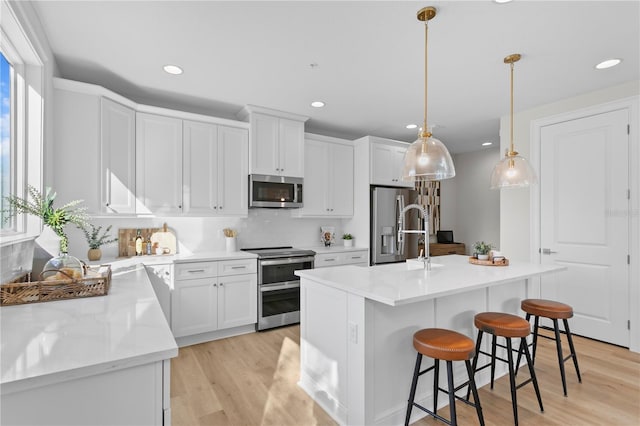 kitchen featuring appliances with stainless steel finishes, decorative light fixtures, light hardwood / wood-style flooring, white cabinets, and an island with sink