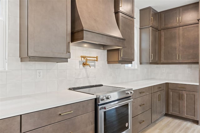kitchen with decorative backsplash, stainless steel range with electric stovetop, custom exhaust hood, light countertops, and light wood-style flooring