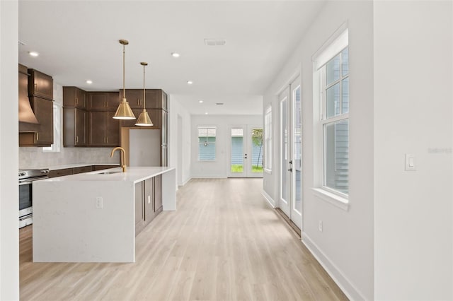 kitchen with sink, an island with sink, hanging light fixtures, wall chimney exhaust hood, and stainless steel range