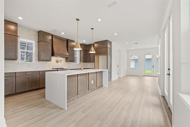 kitchen with a kitchen island with sink, decorative light fixtures, custom exhaust hood, light hardwood / wood-style floors, and tasteful backsplash