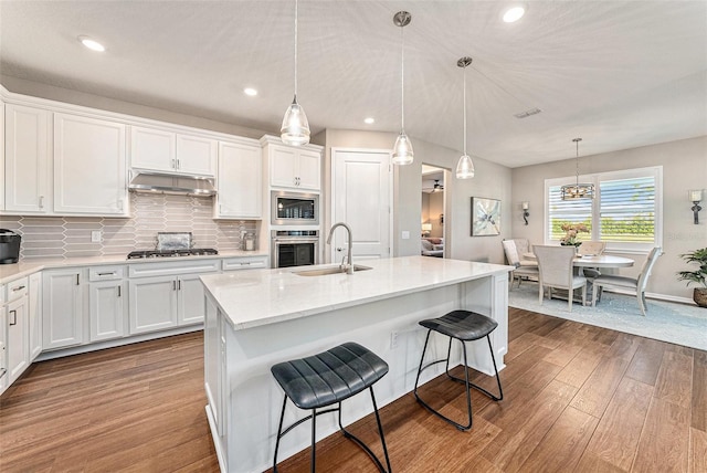 kitchen featuring appliances with stainless steel finishes, hanging light fixtures, light hardwood / wood-style floors, white cabinets, and a center island with sink