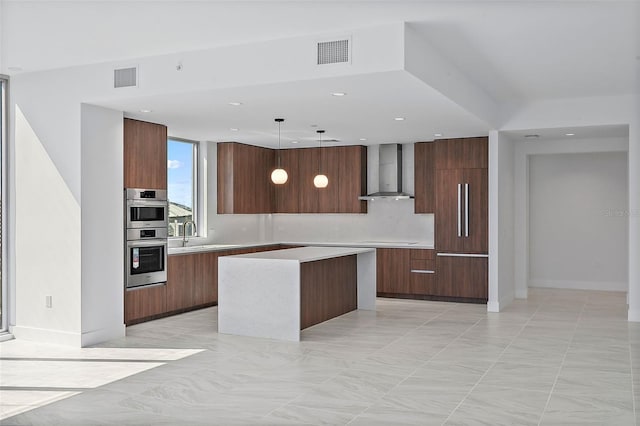 kitchen with wall chimney range hood, a kitchen island, backsplash, stainless steel double oven, and decorative light fixtures