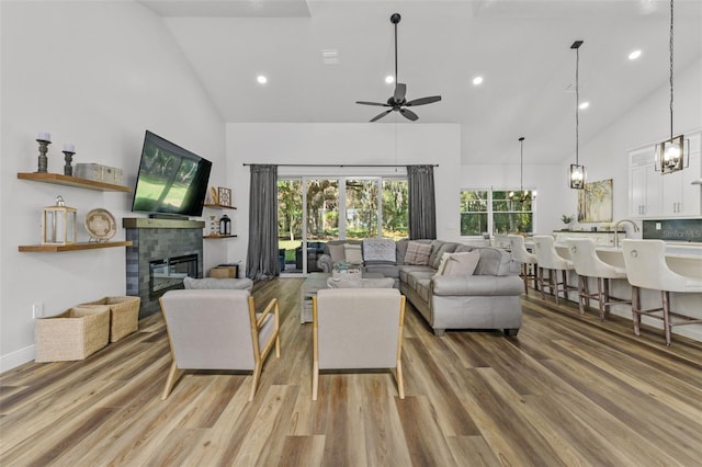 living room with a tiled fireplace, ceiling fan with notable chandelier, wood-type flooring, and high vaulted ceiling