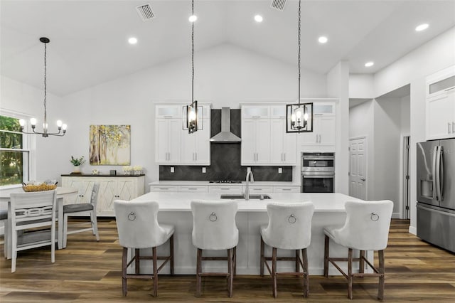 kitchen with wall chimney range hood, tasteful backsplash, a center island with sink, white cabinets, and appliances with stainless steel finishes