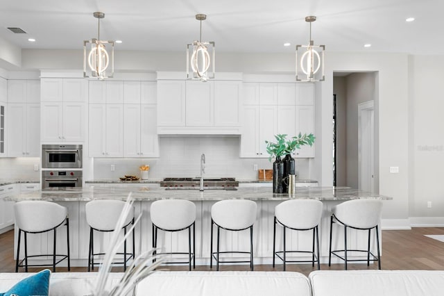 kitchen with double oven, hanging light fixtures, and a center island with sink