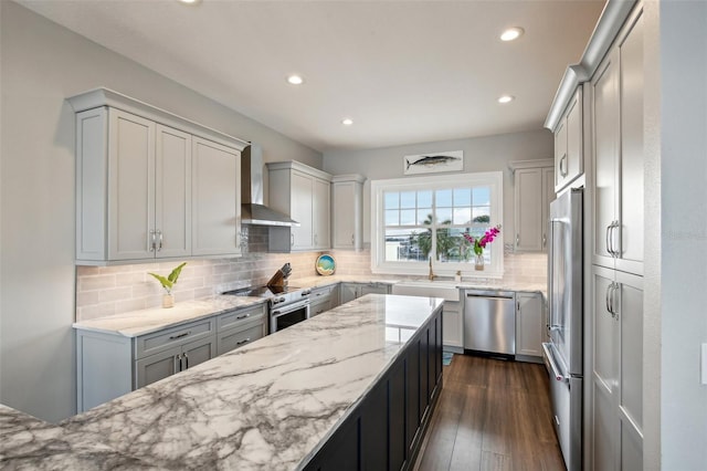 kitchen with wall chimney range hood, dark wood-type flooring, stainless steel appliances, light stone counters, and tasteful backsplash