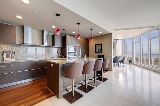 kitchen with wall chimney range hood, stainless steel microwave, light wood-type flooring, decorative light fixtures, and a kitchen island with sink