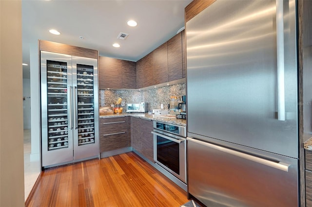 kitchen with light stone countertops, appliances with stainless steel finishes, dark brown cabinets, wine cooler, and light hardwood / wood-style flooring