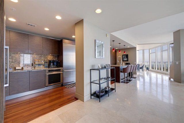 kitchen with oven, light wood-type flooring, dark brown cabinets, hanging light fixtures, and decorative backsplash