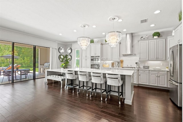 kitchen featuring wall chimney range hood, appliances with stainless steel finishes, dark hardwood / wood-style floors, decorative light fixtures, and a center island with sink