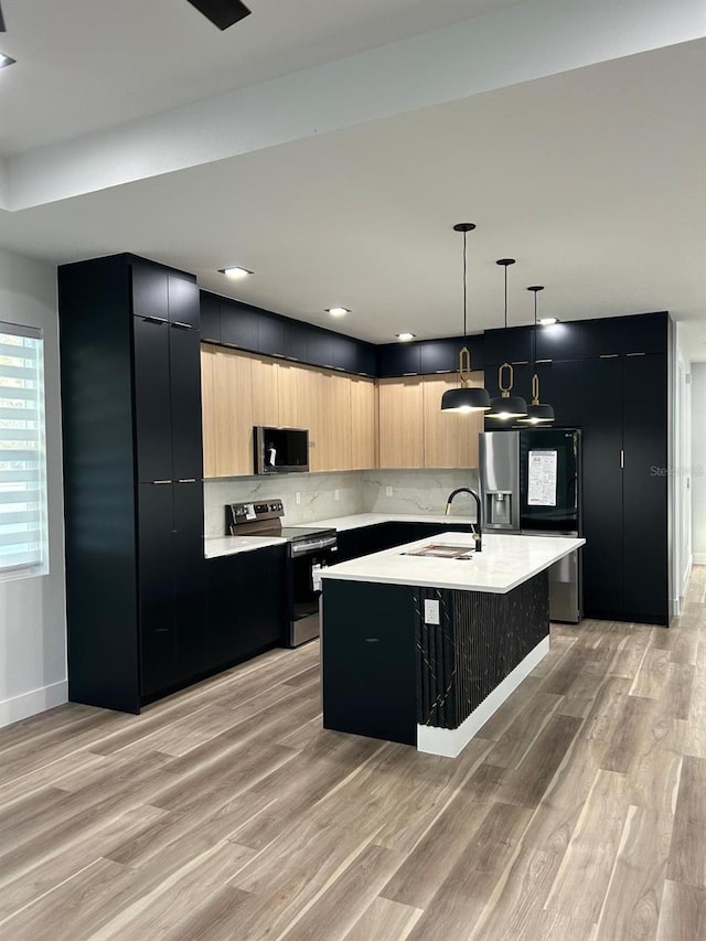 kitchen with light hardwood / wood-style floors, stainless steel appliances, an island with sink, pendant lighting, and light brown cabinetry