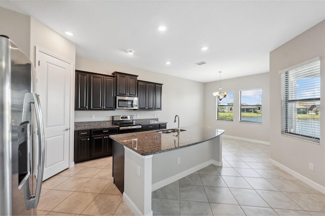 kitchen with a kitchen island with sink, dark stone countertops, decorative light fixtures, light tile patterned floors, and appliances with stainless steel finishes