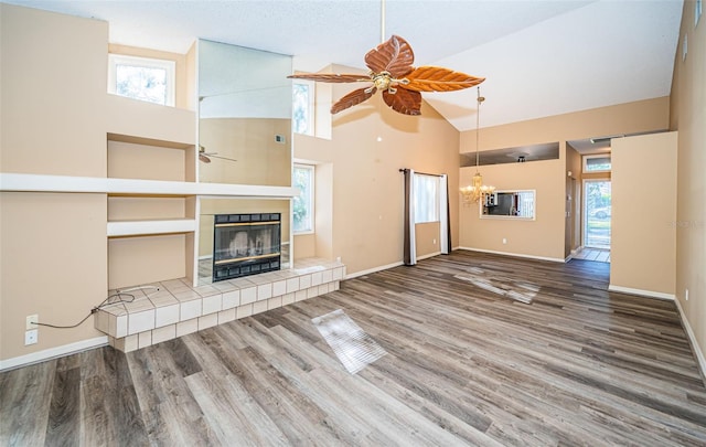 unfurnished living room with baseboards, wood finished floors, a tiled fireplace, high vaulted ceiling, and ceiling fan with notable chandelier
