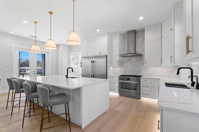 kitchen featuring premium appliances, wall chimney range hood, hanging light fixtures, white cabinets, and a kitchen island with sink