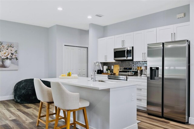 kitchen featuring a kitchen island with sink, stainless steel appliances, decorative backsplash, white cabinets, and light wood-type flooring