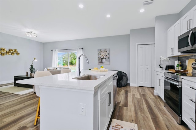 kitchen with stainless steel appliances, a kitchen island with sink, sink, white cabinets, and light hardwood / wood-style floors