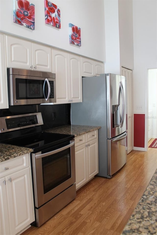 kitchen with white cabinets, appliances with stainless steel finishes, dark stone counters, a high ceiling, and light hardwood / wood-style floors