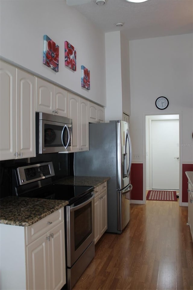 kitchen with white cabinets, dark hardwood / wood-style flooring, appliances with stainless steel finishes, dark stone countertops, and a high ceiling