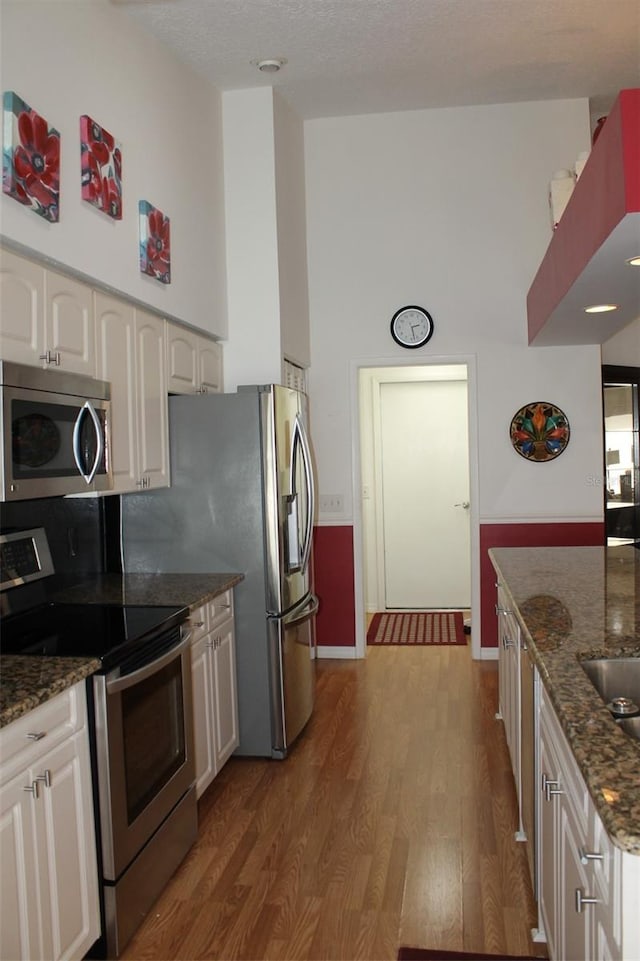 kitchen with appliances with stainless steel finishes, white cabinetry, hardwood / wood-style flooring, dark stone countertops, and a towering ceiling