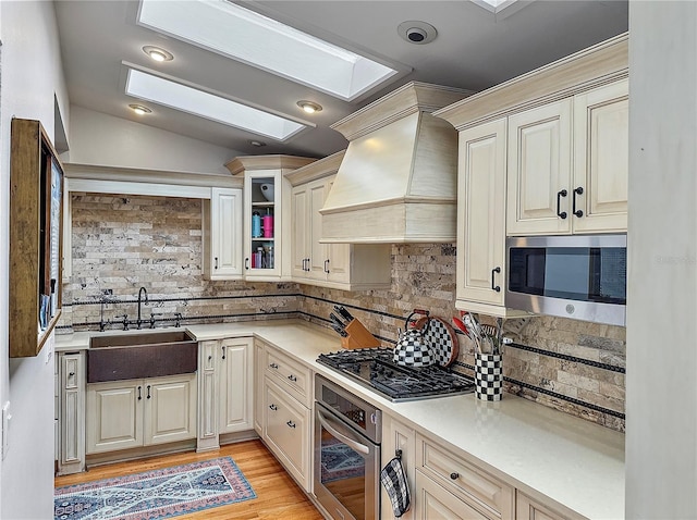 kitchen featuring glass insert cabinets, appliances with stainless steel finishes, light countertops, premium range hood, and a sink