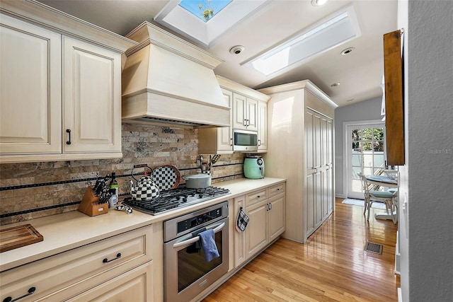 kitchen featuring light countertops, appliances with stainless steel finishes, light wood-style floors, lofted ceiling with skylight, and premium range hood