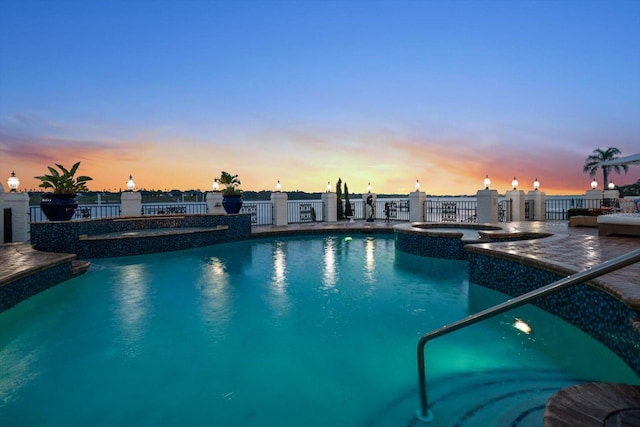 pool at dusk featuring a pool with connected hot tub and fence