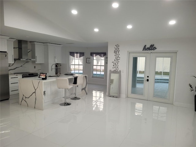 kitchen with a kitchen bar, french doors, a center island with sink, wall chimney range hood, and white cabinetry