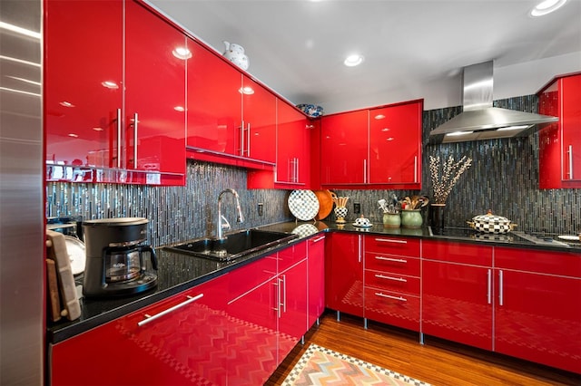 kitchen with wall chimney range hood, red cabinets, tasteful backsplash, wood finished floors, and a sink