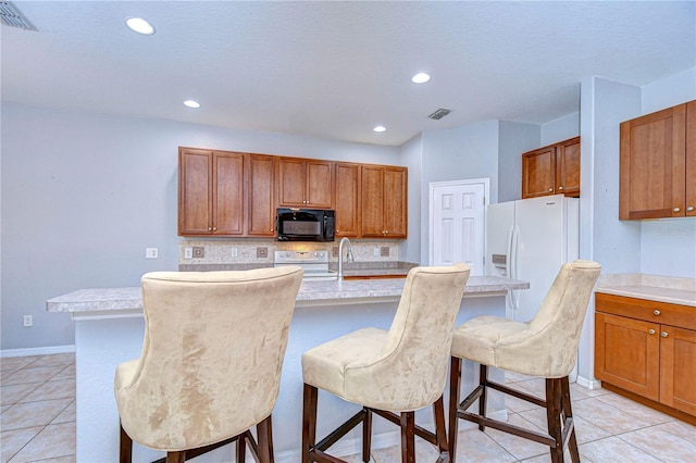 kitchen featuring tasteful backsplash, a center island with sink, a kitchen breakfast bar, light tile patterned flooring, and white appliances