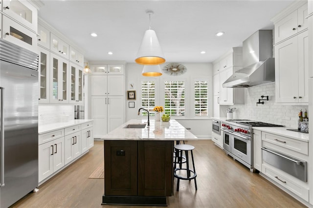 kitchen featuring wall chimney exhaust hood, an island with sink, high end appliances, sink, and white cabinets