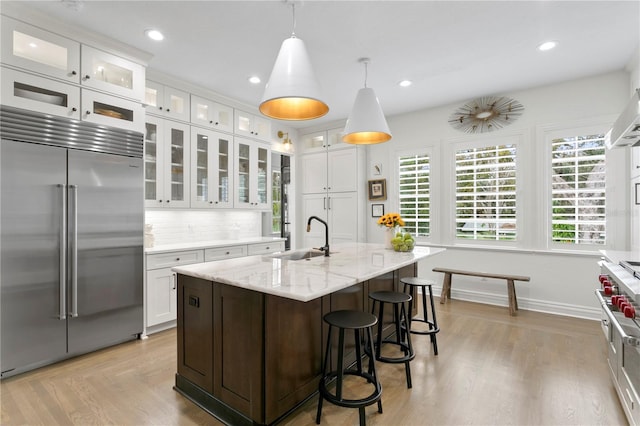 kitchen featuring sink, built in fridge, hanging light fixtures, white cabinets, and a center island with sink