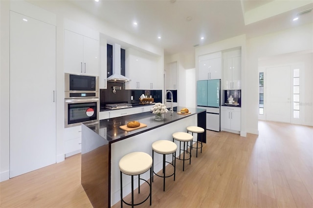 kitchen with appliances with stainless steel finishes, wall chimney range hood, light wood-type flooring, white cabinetry, and a center island with sink