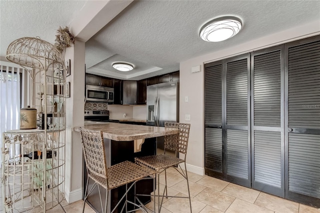 kitchen with a kitchen breakfast bar, dark brown cabinets, a textured ceiling, stainless steel appliances, and light tile patterned floors