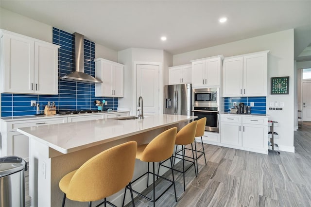 kitchen featuring a center island with sink, white cabinets, a breakfast bar area, wall chimney exhaust hood, and stainless steel appliances