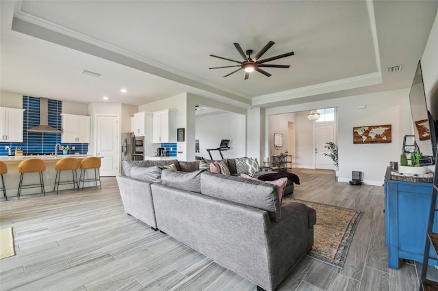 living room featuring a raised ceiling, sink, crown molding, ceiling fan, and light wood-type flooring