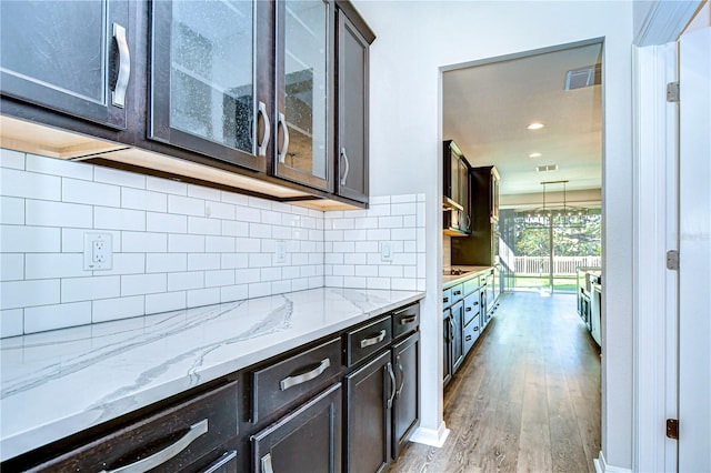 kitchen featuring decorative backsplash, hardwood / wood-style floors, dark brown cabinetry, an inviting chandelier, and light stone counters