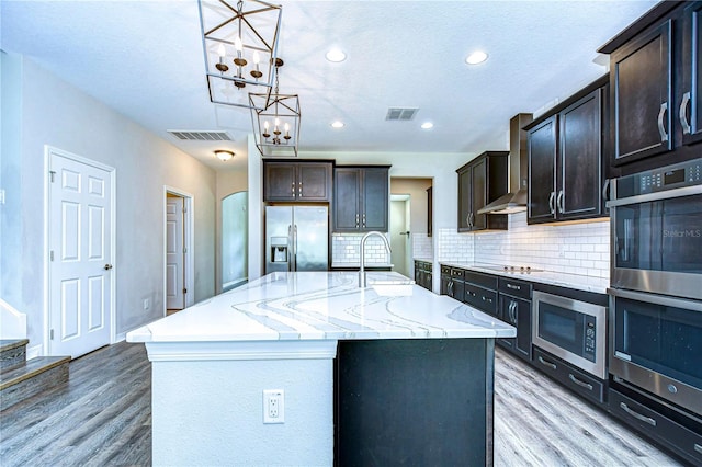 kitchen featuring wall chimney exhaust hood, stainless steel appliances, a center island with sink, sink, and light hardwood / wood-style floors