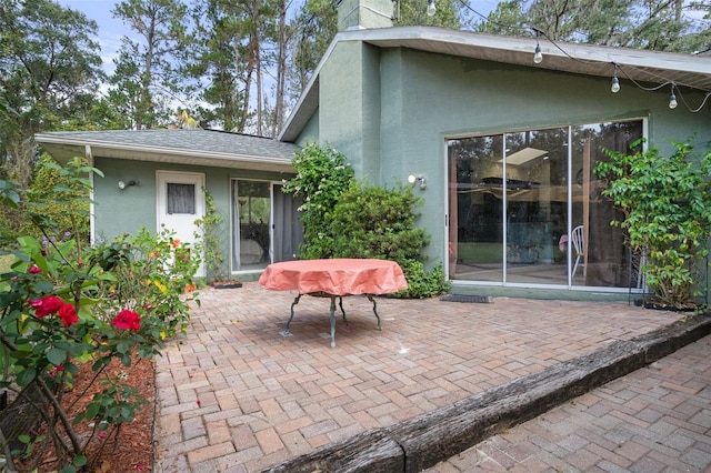 rear view of property with a shingled roof, a chimney, stucco siding, and a patio