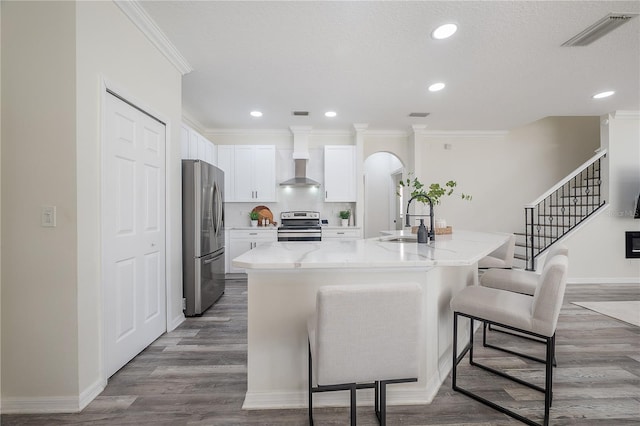 kitchen with wall chimney range hood, an island with sink, stainless steel appliances, sink, and white cabinetry