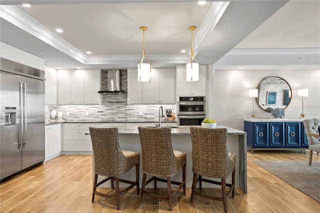 kitchen featuring appliances with stainless steel finishes, blue cabinets, hanging light fixtures, white cabinetry, and wall chimney exhaust hood