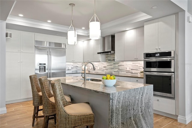 kitchen with white cabinets, a center island with sink, pendant lighting, wall chimney exhaust hood, and stainless steel appliances