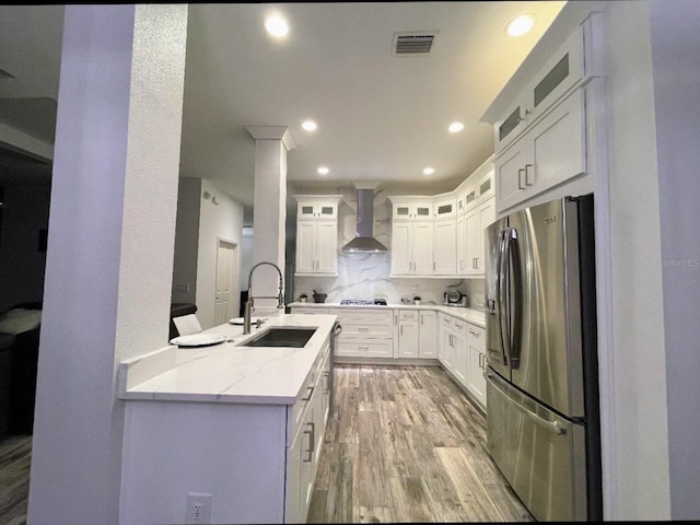 kitchen with wall chimney range hood, sink, stainless steel refrigerator with ice dispenser, light stone counters, and white cabinetry