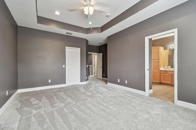 unfurnished bedroom with a raised ceiling, ensuite bathroom, ceiling fan, and light colored carpet