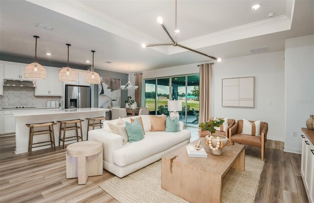 living room with crown molding, light wood-type flooring, an inviting chandelier, sink, and a tray ceiling