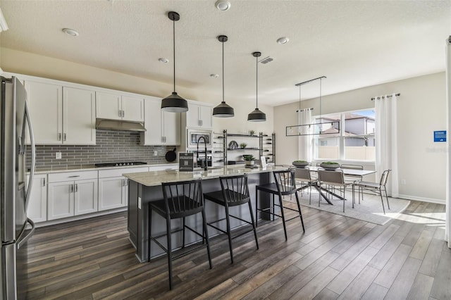 kitchen with hanging light fixtures, a center island with sink, appliances with stainless steel finishes, white cabinetry, and dark wood-type flooring