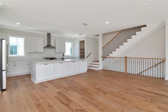 kitchen featuring an island with sink, wall chimney range hood, light wood-type flooring, light countertops, and white cabinets