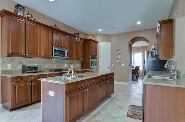 kitchen featuring decorative backsplash, appliances with stainless steel finishes, light stone countertops, sink, and an island with sink