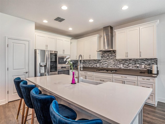 kitchen with wall chimney range hood, dark hardwood / wood-style flooring, stainless steel appliances, white cabinets, and a kitchen island with sink