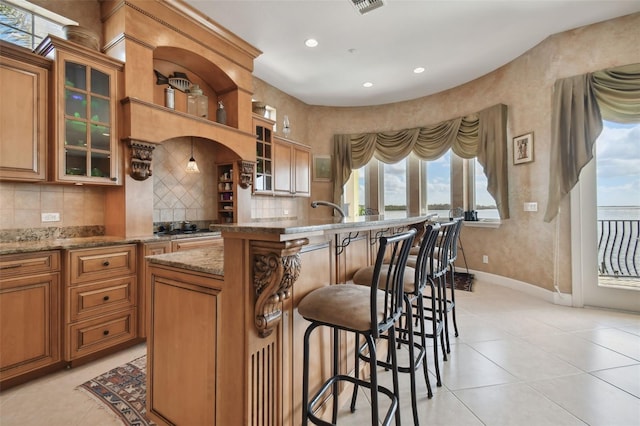 kitchen with a kitchen bar, a center island with sink, tasteful backsplash, light tile patterned flooring, and light stone counters