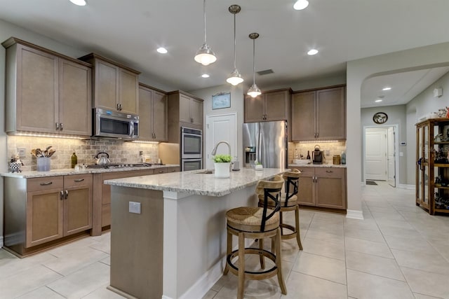 kitchen featuring stainless steel appliances, light stone counters, tasteful backsplash, a kitchen island with sink, and pendant lighting